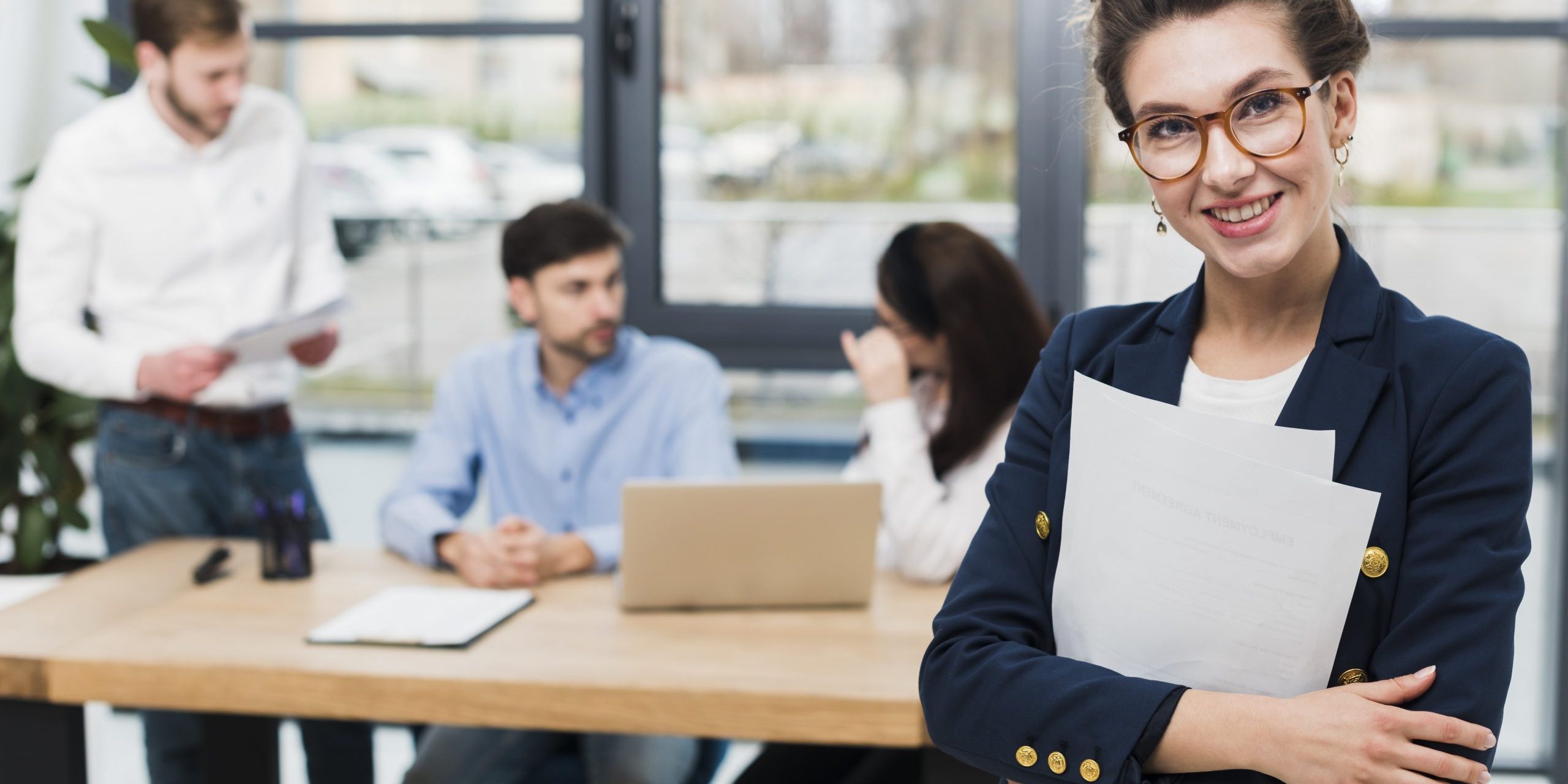Woman in business attire with people sitting at desk in the background illustrates blog "Will a Background Check Show All My Jobs?"