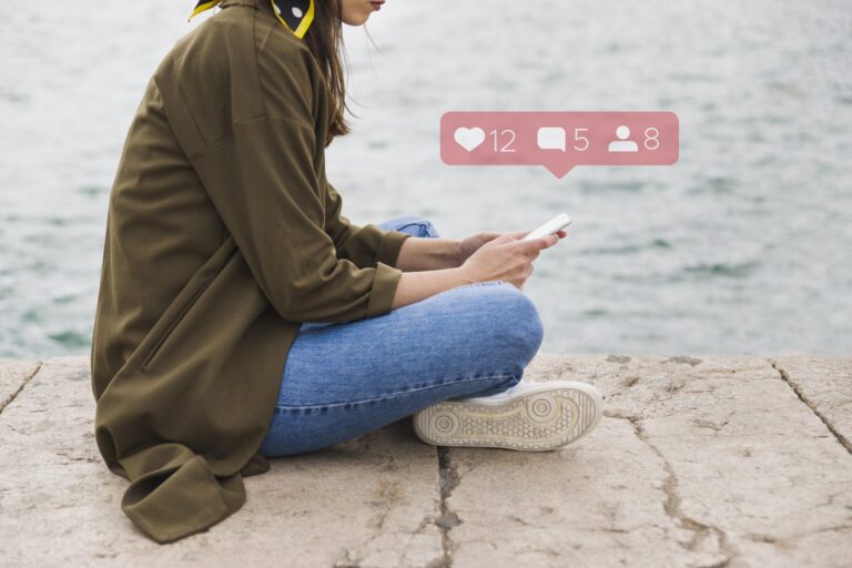 Woman wearing jeans and a green jacket sitting on the floor holding smartphone with social media icons hovering above it.